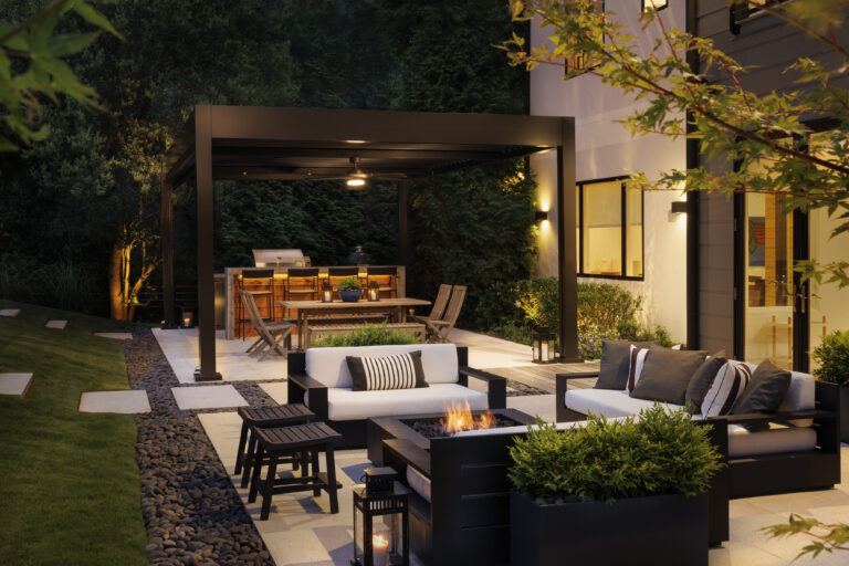 Modern backyard patio with black aluminum pergola, outdoor kitchen bar, fire pit seating, and warm landscape lighting at night